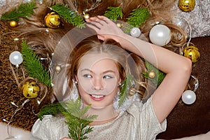 Top view of a blonde young girl lying against a background of golden and white Christmas balls and fir branches in the studio. New