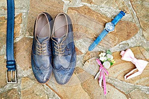 Top view of blue leather groom shoes, watches, belt, boutonniere, pink bowtie on brown natural stone. Groom wedding accessories.