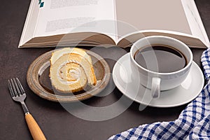 Top view of a book with roll cakes on a plate, a white coffee cup, and a cloth on dark gray stone background.
