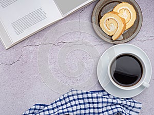 Top view of a book with roll cakes on a plate, a white coffee cup, and a cloth on a gray stone background. Space for text.