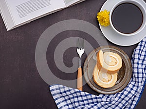 Top view of a book with roll cakes on a plate, a white coffee cup, and a cloth placed on dark gray stone background.