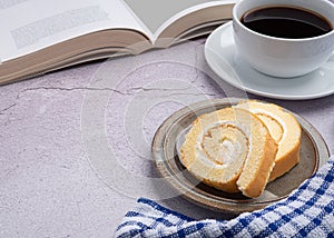 Top view of a book with roll cakes on a plate, a white coffee cup, and a cloth on a table.