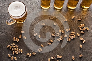 Top view of bottles and mug of light beer near scattered pistachios on brown surface.