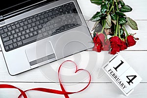 Top view of bouquet of red roses, wood calendar february 14, ribbon shaped as heart and open laptop computer on white table.