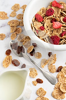 Top view of bowl with cereals, red berries, spoon and jug with milk, on white marble table