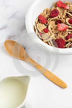 Top view of bowl with cereals, red berries, wooden spoon and jug with milk, on white marble table