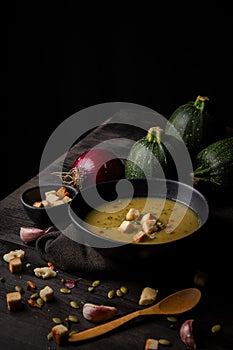 Top view of a bowl of homemade zucchini cream with pumpkin seeds, bread and paprika, on dark table with cloth,