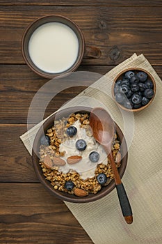 Top view of a bowl of muesli, a glass of milk and a bowl of blueberries on a wooden table. Flat lay