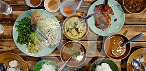 Top view bowl of spicy fish curry sauce and other food on wooden table with Thai rice noodle, vegetable, grilled chicken