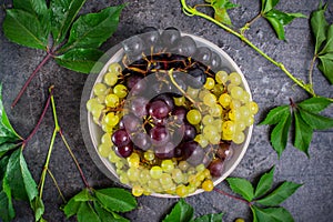 Top view bowl of various grapes: red, white and black berries and green leaves with water drops on the dark concrete background. S