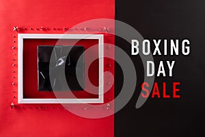 Top view of Boxing day Sale text on white picture frame with black gift box and Christmas ball and berries on red background.