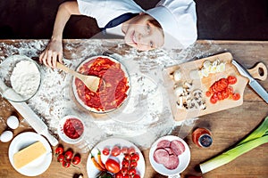 Top view of boy making pizza with pizza ingredients, tomatoes, salami and mushrooms