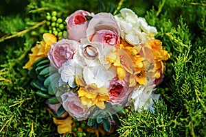 Top view of bridal bouquet with white, yellow and rose flowers with wedding rings on it lying on grass in summer wedding day.