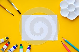 Top view of brushes, color tubes, pencils, palette and white paper on yellow background, copy space, flat lay