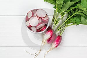 Top view of a bunch of radishes and a bowl of radish slices on a white wooden table