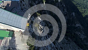 Top view of the cable car station with a cabin approaching to it and people walking near the cliff edge. Shot. Aerial of