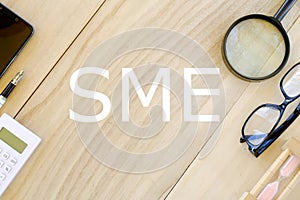 Top view of calculator,pen,mobile phone,magnifying glass,sunglasses and sand clock on wooden background written with SME Small an