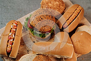 Top view of a cheeseburger and hot dogs with breads next to them on a wooden board.