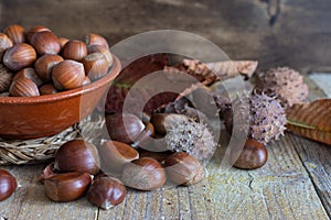 Top view of chestnuts with leaves and ceramic bowl with hazelnuts in the background, on weathered wooden background