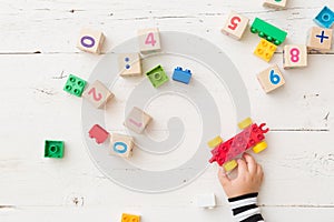 Top view on child`s hand playing with wooden cubes with numbers and colorful toy bricks on white wooden background. Baby with toy