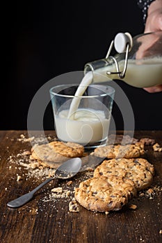 Top view of chocolate chip cookies, crumbs, spoon and hand with bottle pouring milk into glass cup, on wooden table and black back