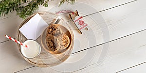 Top view Christmas composition. Glass milk, cookies, Santa's sleigh, empty note, white wooden table.