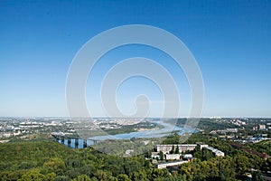 Top view of the city, the bend of the river, the bridge, residential buildings and a green array of trees. Blue sky.