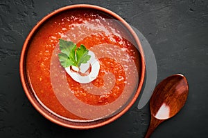 Top view close-up of a brown bowl of tomato soup with parsley and milk cream and a wooden spoon on a dark background