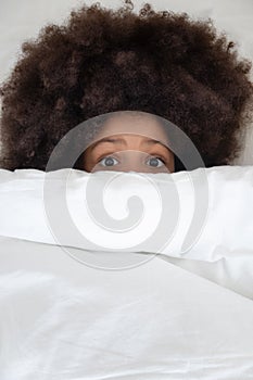 Top view close up scared black kid hiding under duvet.