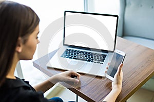 Top view,close-up of smartphone with blank screen in hands of young woman sitting at round wooden table with laptop