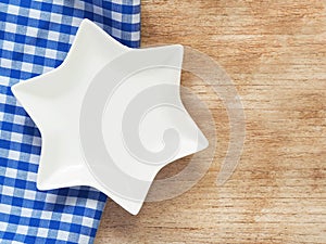 Top view, close-up of star-shape white plate on blue and white checkered fabric or napkin on wooden table background.