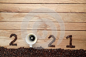 Top view of coffee beans and hot fresh coffee in a white cup with foam and text 2021 for Happy New Year Concept. on wooden table