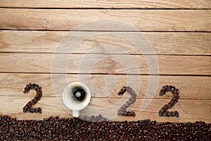 Top view of coffee beans and hot fresh coffee in a white cup with foam and text 2022 for Happy New Year Concept. on wooden table