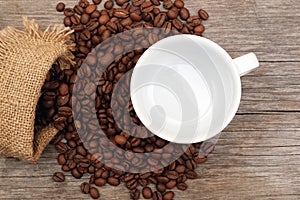 Top view on coffee beans spilled from burlap sack and white empty cup on aged wooden background