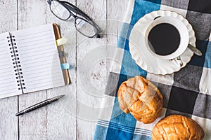 Top view of Coffee cup with bread, tablecloth, notebook, pen and glasses on wooden table background.