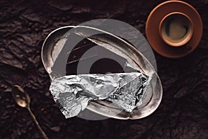 top view of coffee cup, spoon and chocolate in foil on silver trail surrounded by coffee grains on surface covering by black