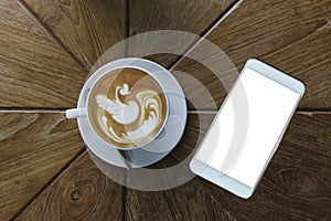 Top view of coffee latte art style in white ceramic cup besides white smart phone with blank white screen