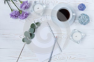 Top view of coffee mug, blank notepad, pencil, flowers and candles on white table. Flat lay
