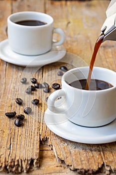 Top view of coffee pot serving in white cup, on rustic wooden table with full cup and coffee beans, selective focus