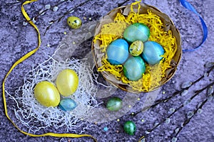 Top view on colored easter eggs in a paper nest and basket and bright ribbons on a dark gray background