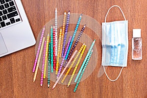 Top view of colored pencils with disinfectant gel, mask and a laptop detail on a wooden surface