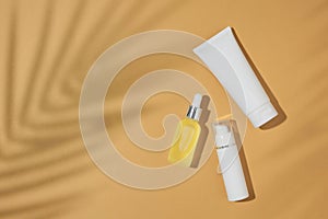 Top view composition made of cosmetic tubes, dropper bottle, pump bottle and tropical leaves shadow on sandy background