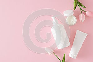 Top view composition made of cosmetic tubes without label, cream jars and spring flowers on pastel pink background