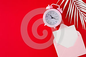 Top view composition made of open envelope with card, white tropical leaves and alarm clock on red background