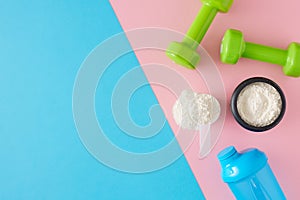 Top view composition of whey protein in jar with shaker and dumbbells on pastel pink and blue background.