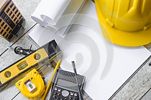 Top view construction tools such as a yellow hard hat, spirit level, measuring tape, folding ruler arrayed against a wooden plank