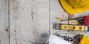 Top view construction tools such as a yellow hard hat, spirit level, measuring tape, folding ruler arrayed against a wooden plank