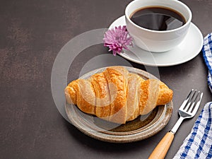 Top view of a croissant on a plate and a white coffee cup with a cloth and cutlery on a dark gray background.