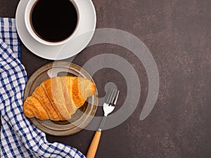 Top view of a croissant on a plate and a white coffee cup with a cloth and cutlery placed on a dark gray background.