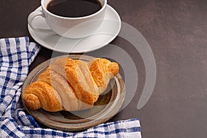 Top view of a croissant on a plate and a white coffee cup with a cloth on a dark gray background.